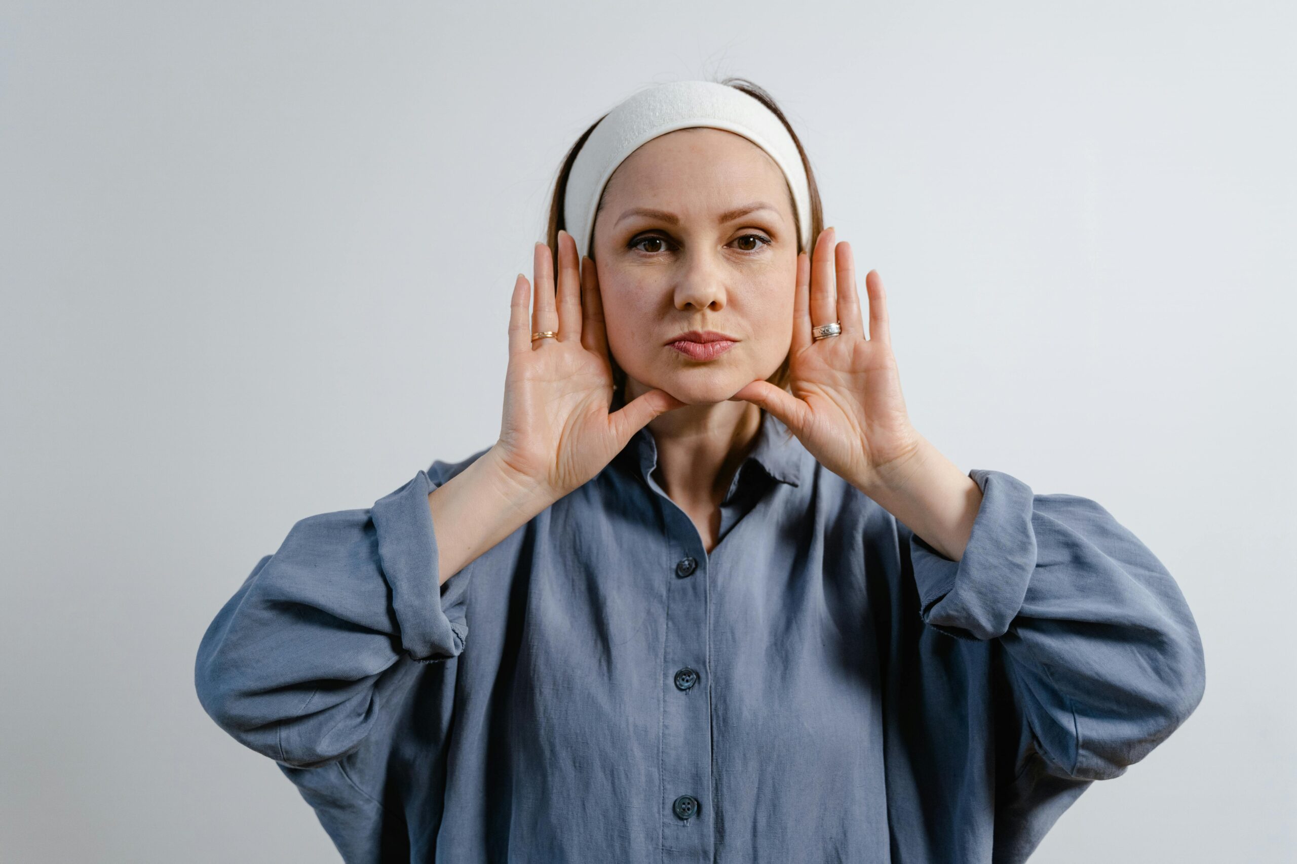 Portrait of a woman in a blue shirt with hands on her face, wearing a headband, on a white background.