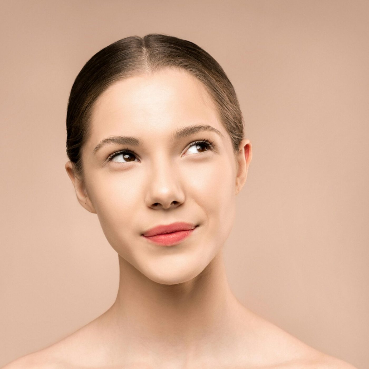 A young woman with radiant skin looks upward against a neutral background, embodying natural beauty.